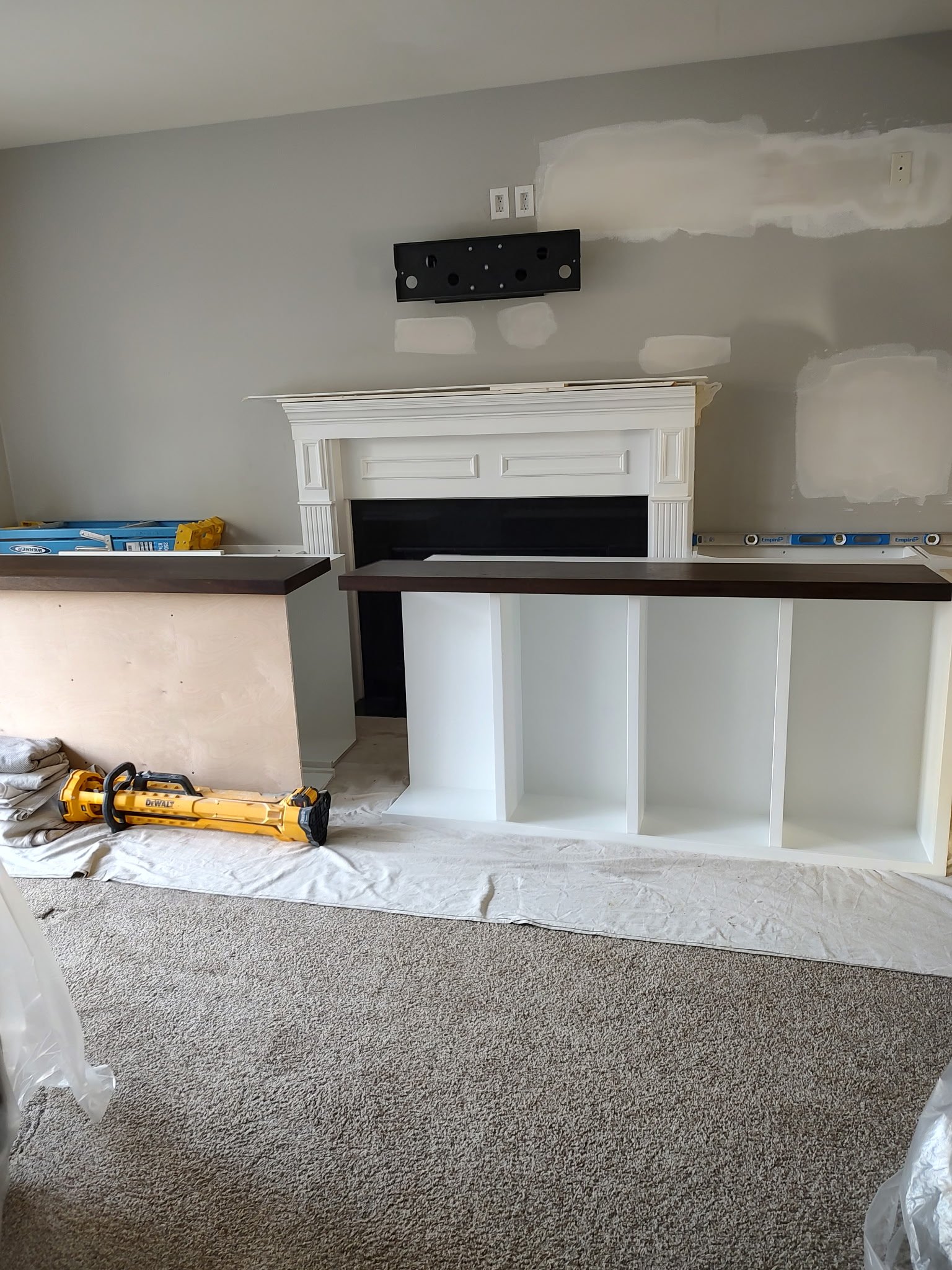 A living room under renovation with a white fireplace, two white shelving units with dark tops, a wall-mounted TV bracket, painting tools, and drop cloths covering the carpet and nearby furniture.