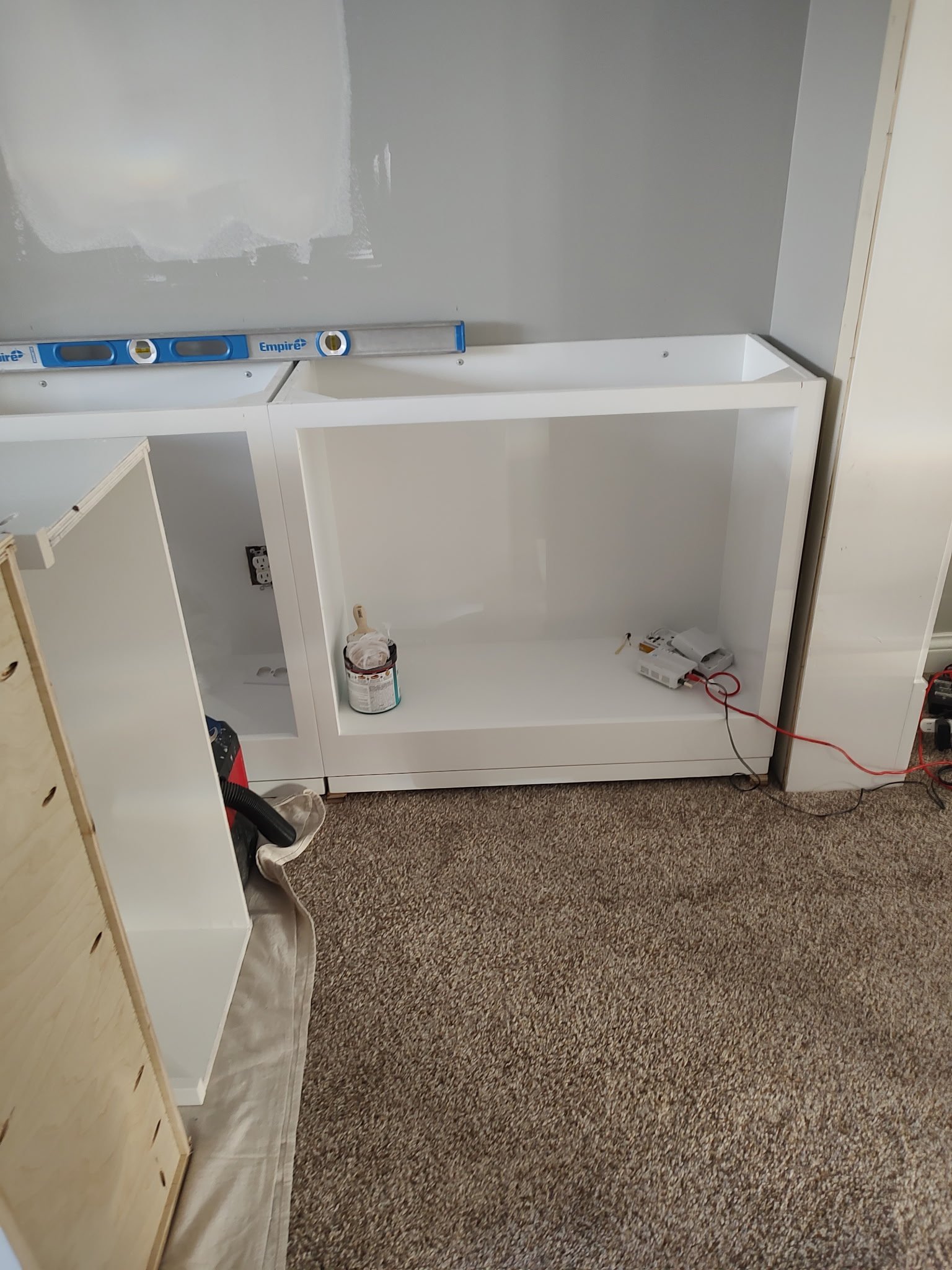 A partially assembled white cabinet sits on a carpeted floor near a gray wall. Tools, a paint can, a level, and electrical cords are nearby, suggesting ongoing home improvement or renovation work.
