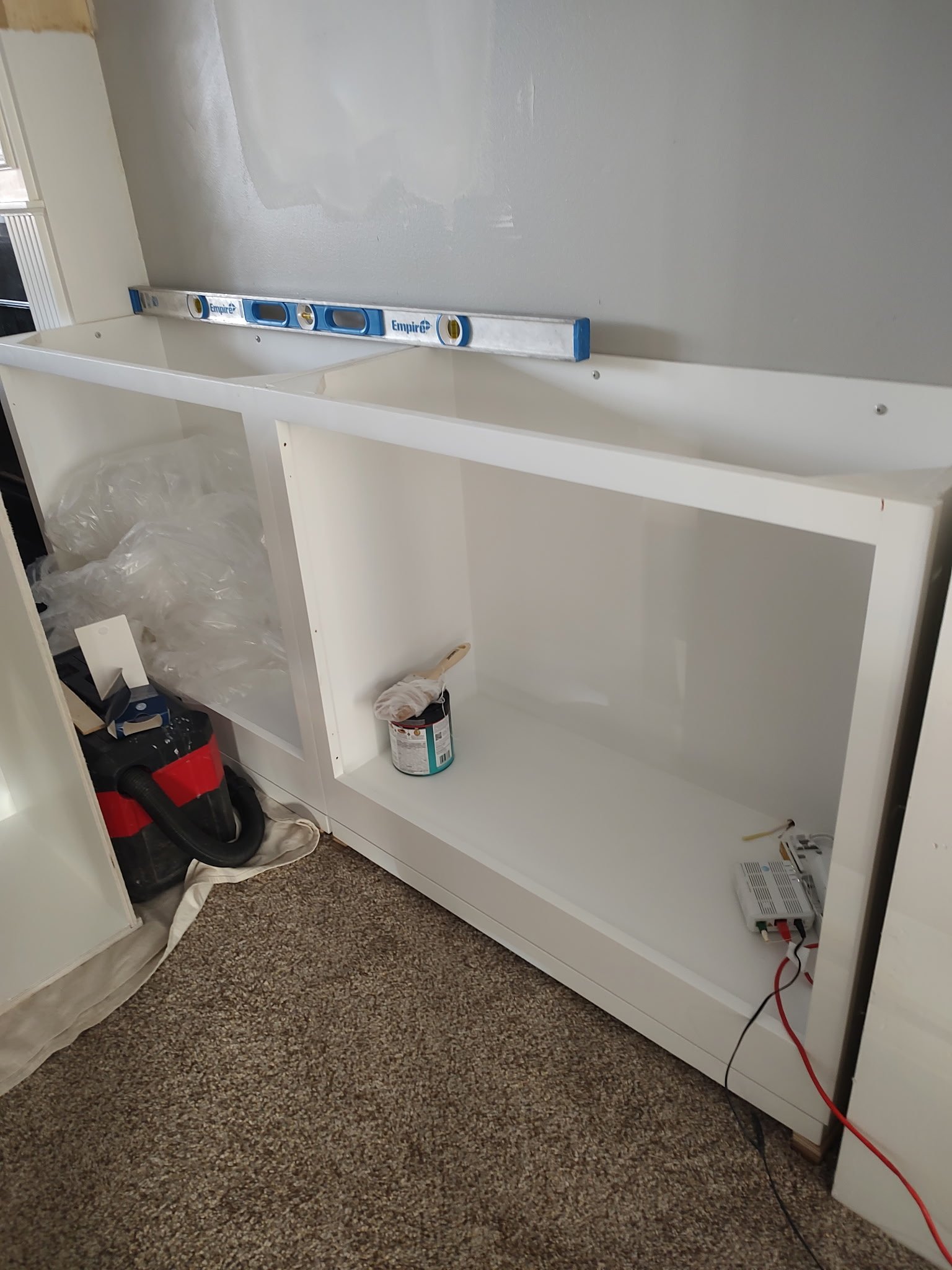 A partially assembled white cabinet sits on a carpeted floor against a gray wall. A level rests on top, and paint supplies, a vacuum, and wires are nearby. Plastic sheeting and a tarp are on the left side.