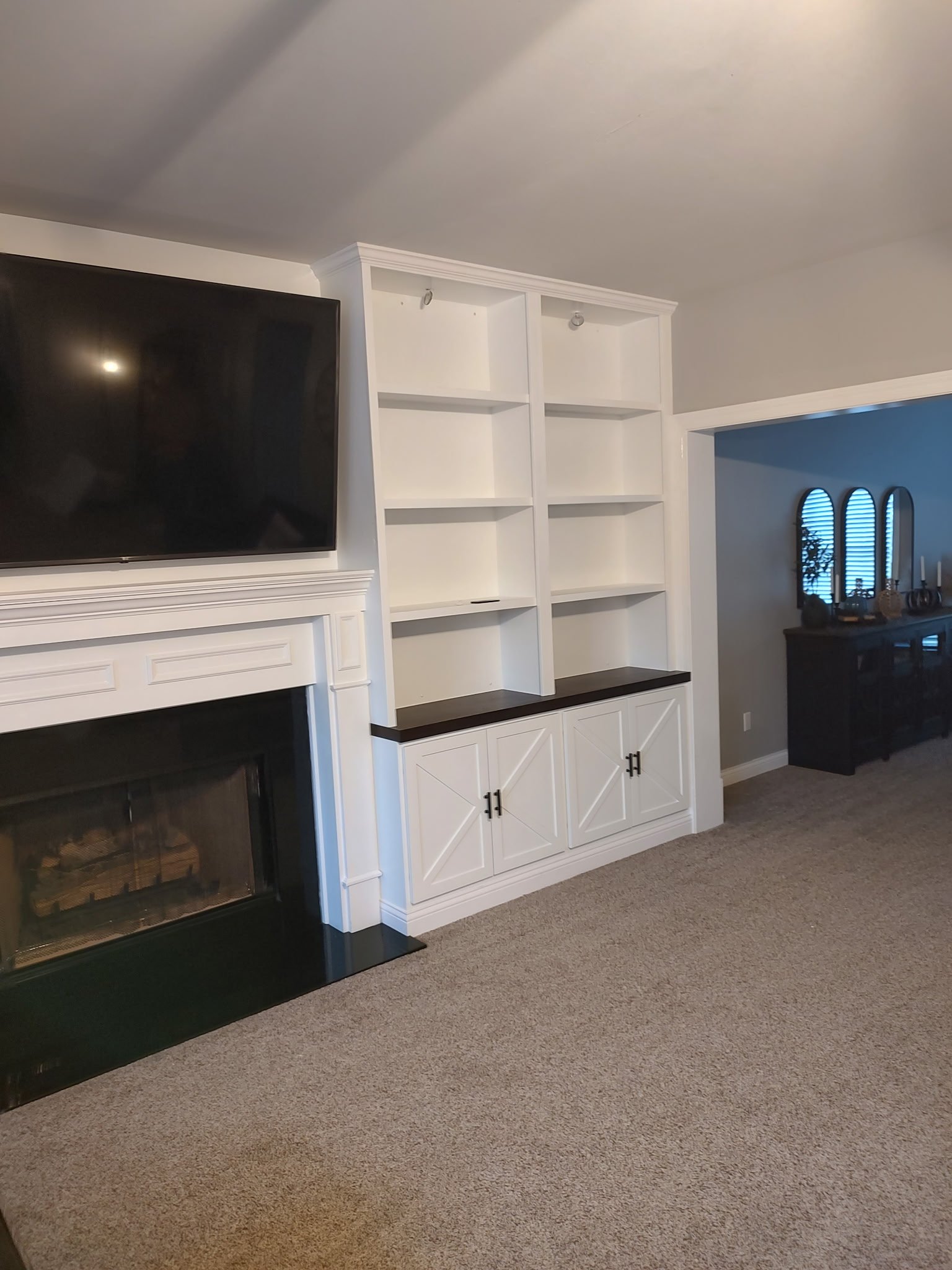 A living room with a wall-mounted TV above a white fireplace, built-in white shelves with cabinets below, and carpeted flooring. An adjoining room with a sideboard and mirrors is visible through an open doorway.