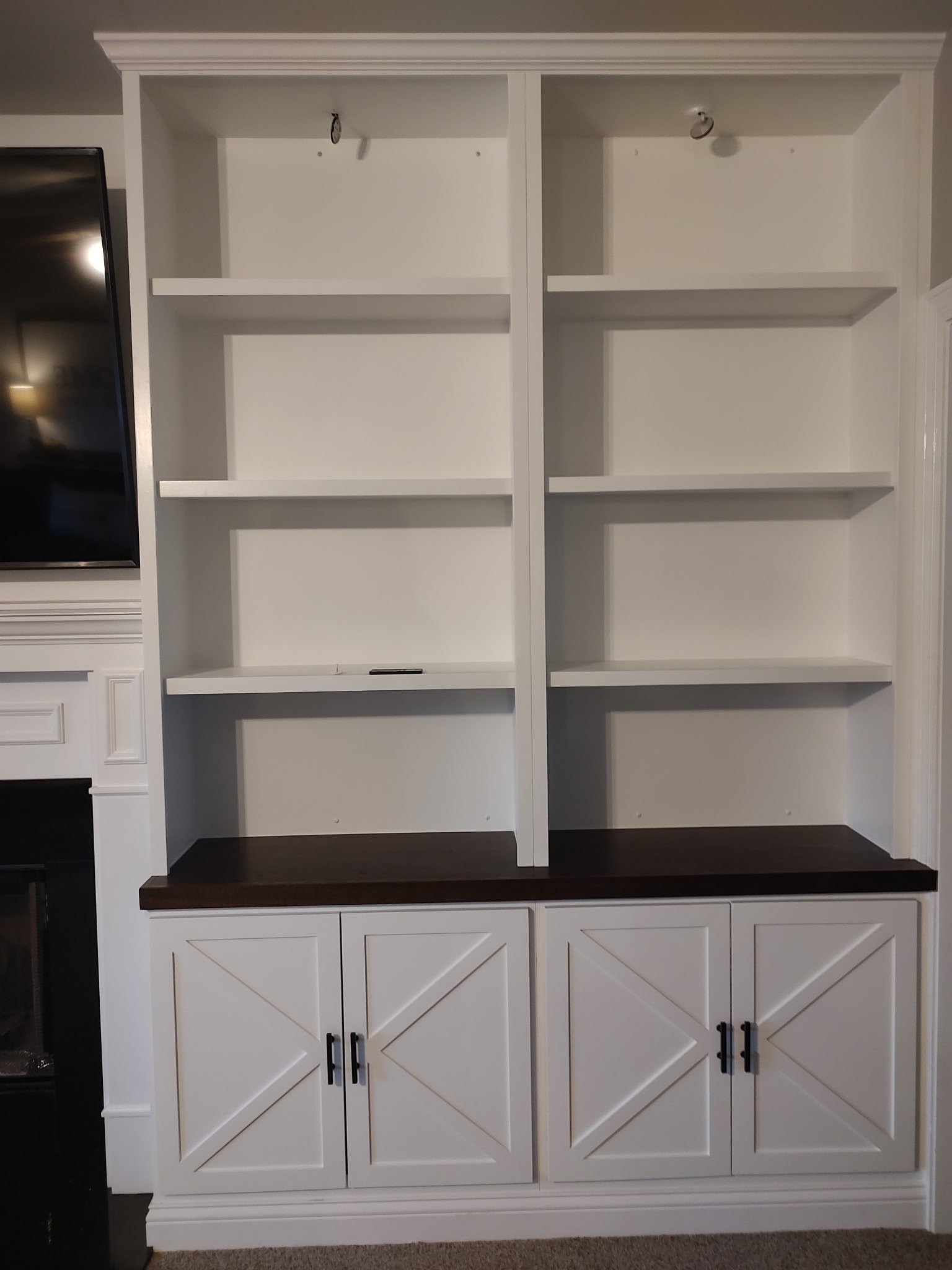 A built-in white shelving unit with four open shelves and four lower cabinets featuring dark handles. The countertop is dark wood, and the unit is next to a fireplace and TV. The shelves are currently empty.
