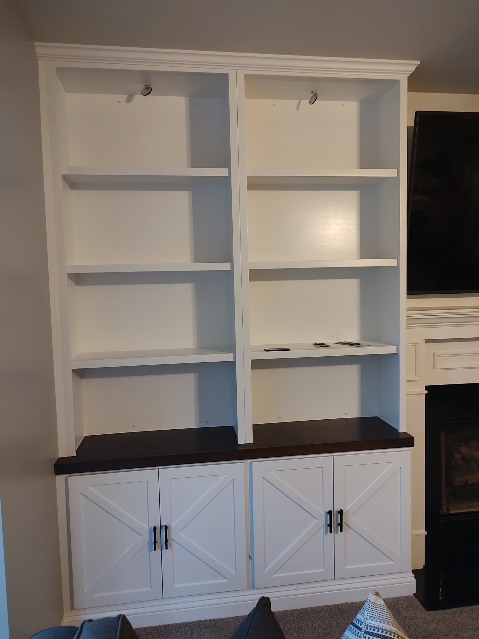 A white built-in shelving unit with four empty upper shelves and two cabinets with X-shaped panel doors below, set against a light-colored wall beside a fireplace.