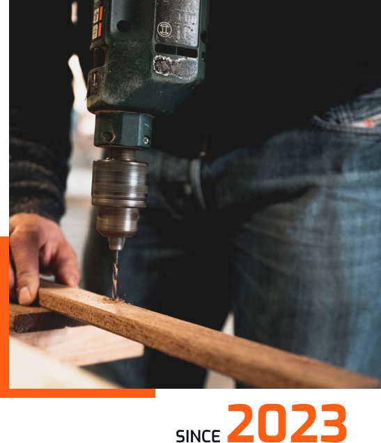 A person uses a handheld electric drill to make a hole in a wooden board. The photo focuses on the drill and the wood, with Since 2023 written at the bottom in orange and black text.