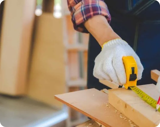 A person wearing a plaid shirt and white glove measures a piece of wood with a yellow tape measure in a woodworking setting.